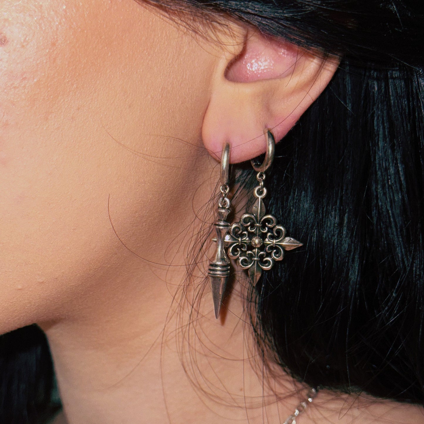 Two fleur-de-lis inspired dangle earrings in oxidized silver-tone worn together on a model with dark hair