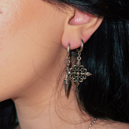 Two fleur-de-lis inspired dangle earrings in oxidized silver-tone worn together on a model with dark hair