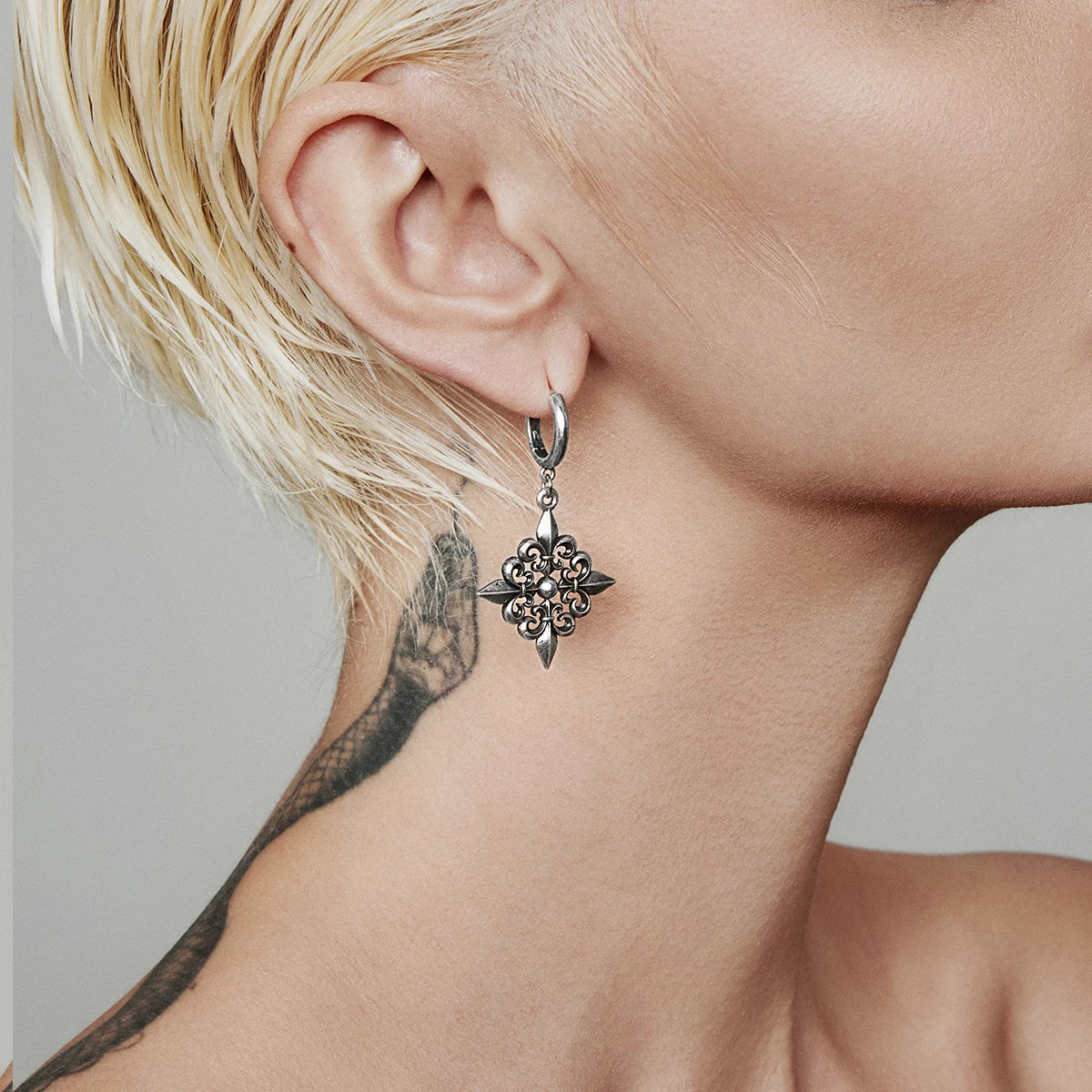 Fleur-de-lis dangle earring in antiqued silver-tone worn on a model with short blonde hair and neck tattoo