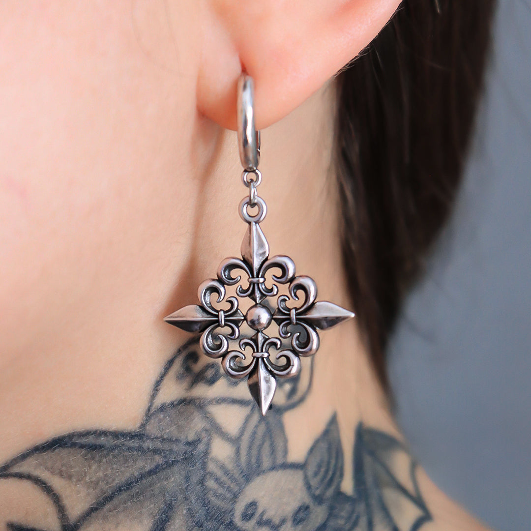 Close-up of a fleur-de-lis dangle earring in dark antiqued silver-tone worn on a model with a tattooed neck