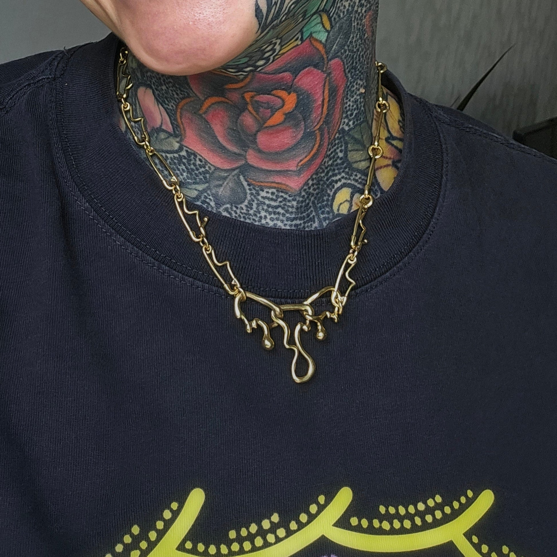 Gold-tone fluid pin necklace worn by a model with a neck tattoo over a dark sweatshirt, close-up