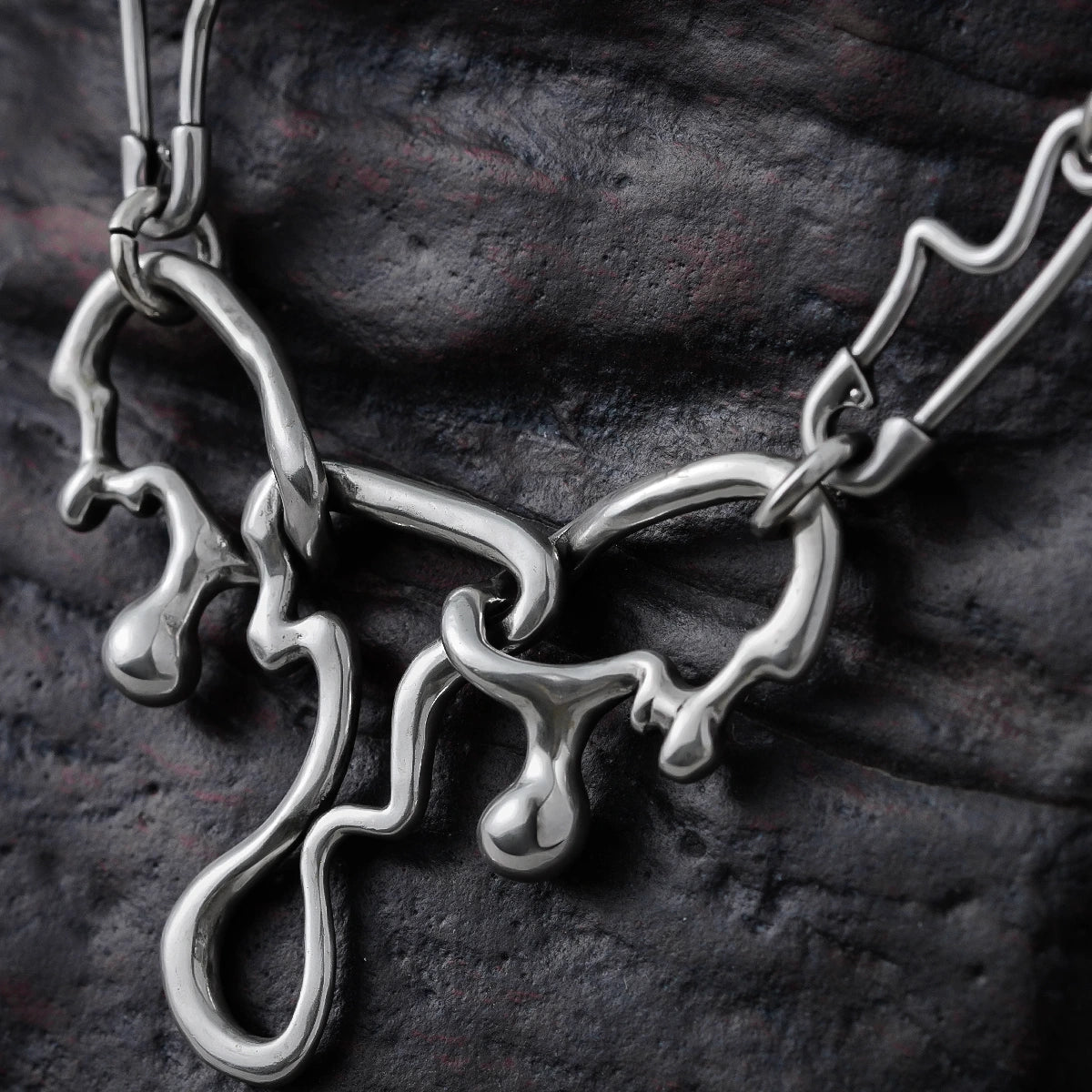 Close-up of a silver-tone fluid pin necklace pendant with dripping abstract links against a dark textured surface