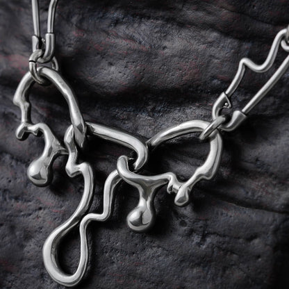 Close-up of a silver-tone fluid pin necklace pendant with dripping abstract links against a dark textured surface