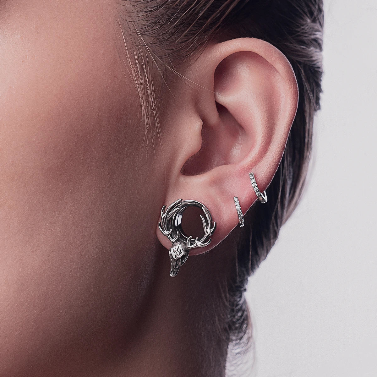 Model wearing a silver-tone deer skull hoop earring with antlers next to two crystal stud earrings