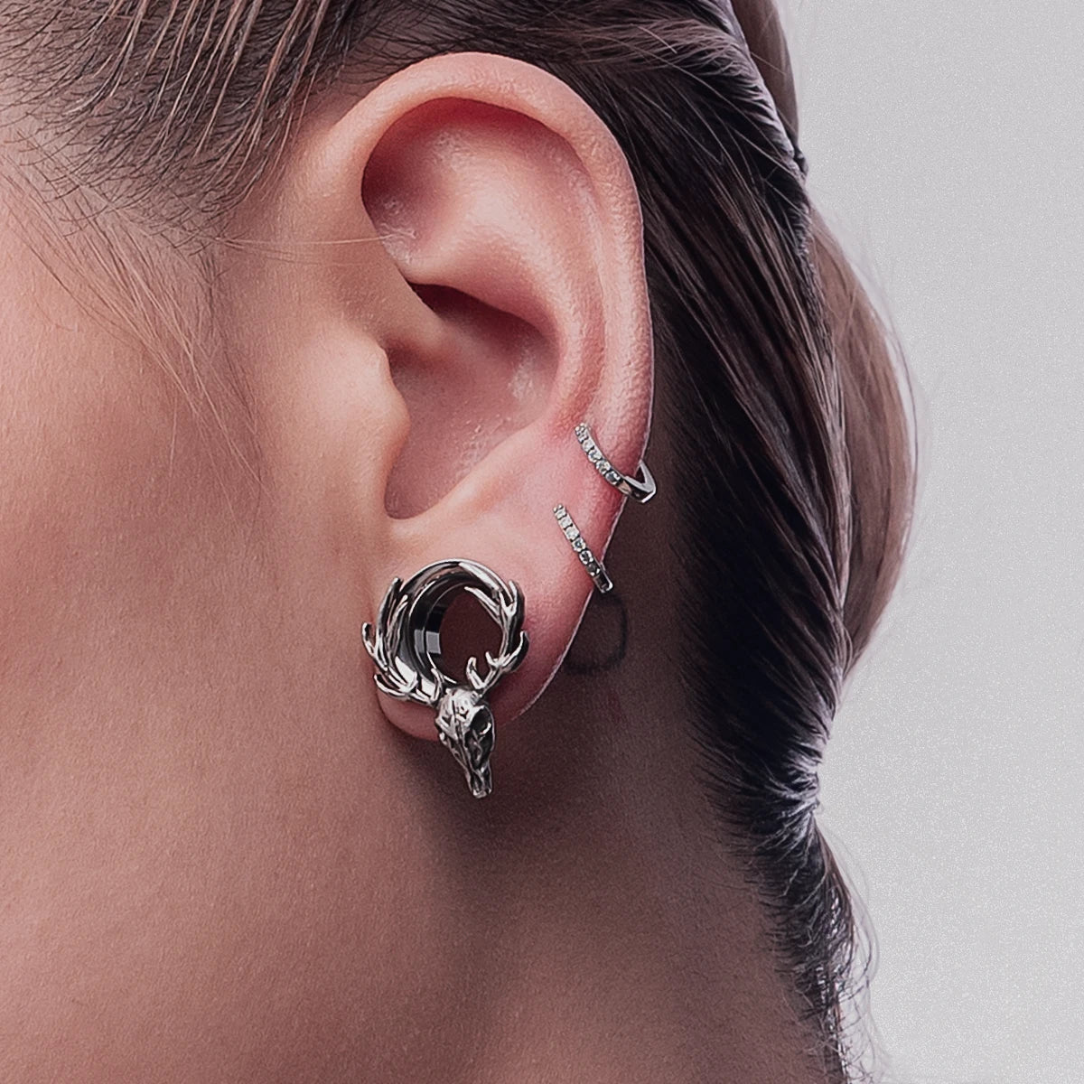 Close view of a silver-tone deer skull hoop earring worn with crystal cartilage hoops