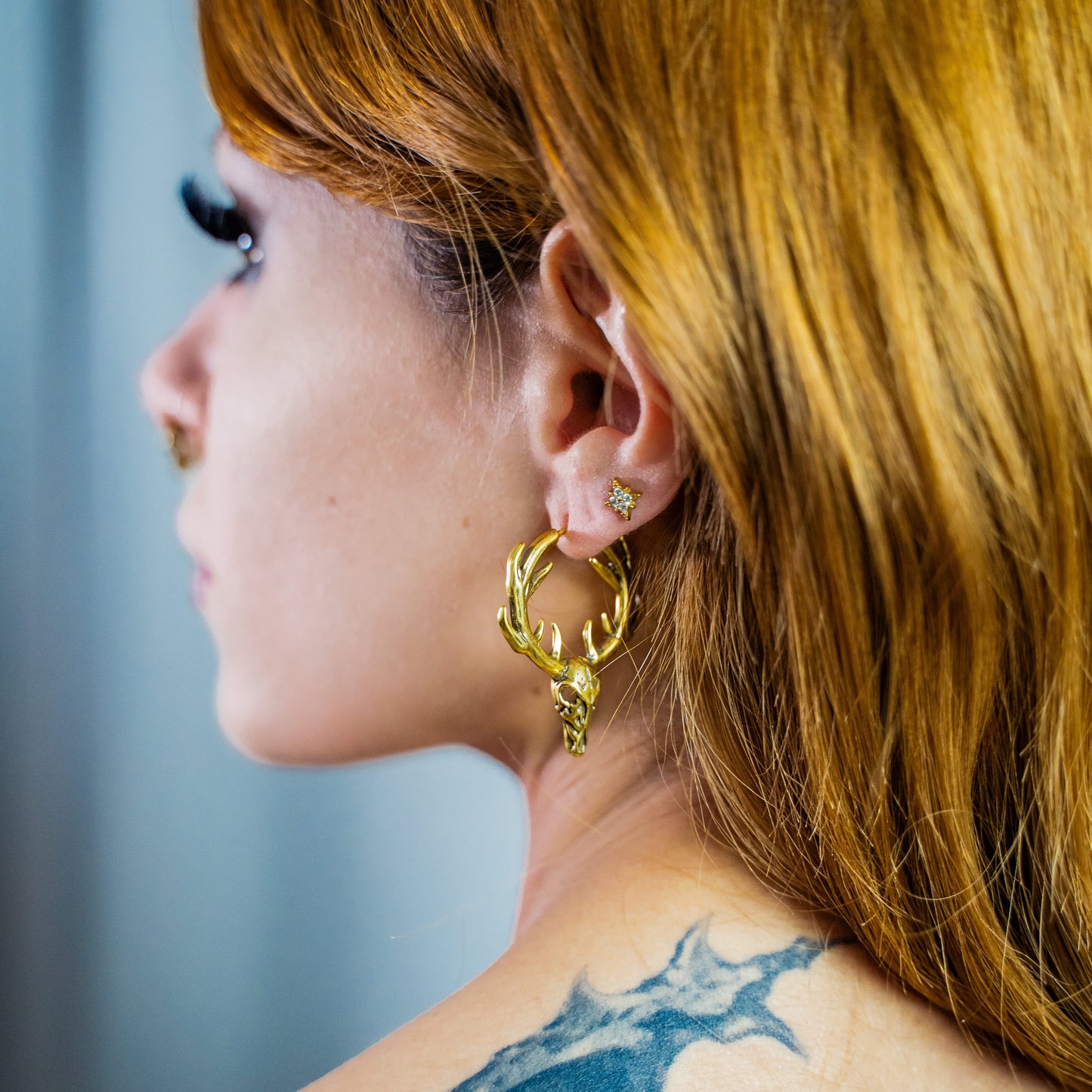 Gold-tone stag skull hoop earring with antlers worn on a model's ear, side profile with tattooed shoulder