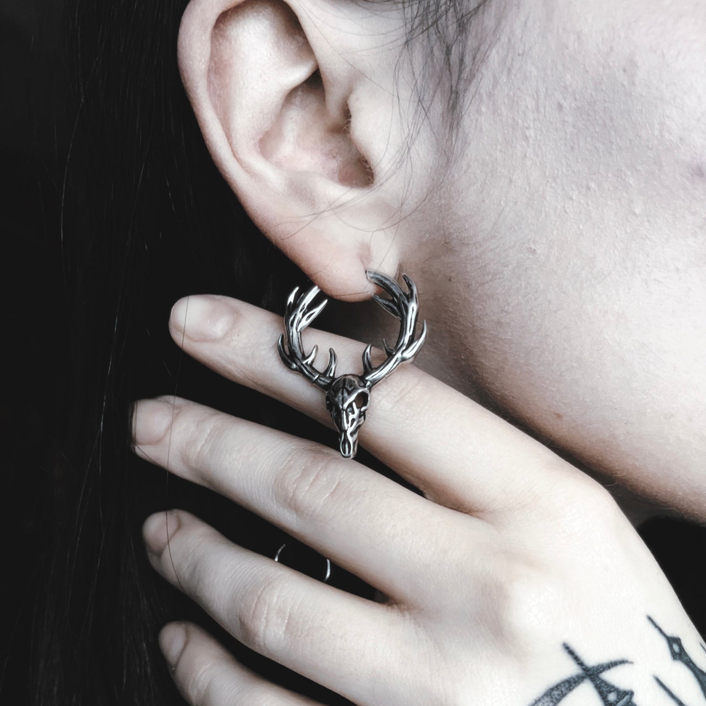 Close-up of a silver-tone stag skull hoop earring with antlers worn on a model's ear, hand resting near the jaw
