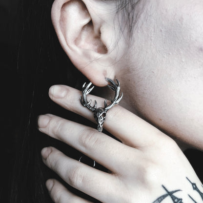Close-up of a silver-tone stag skull hoop earring with antlers worn on a model's ear, hand resting near the jaw