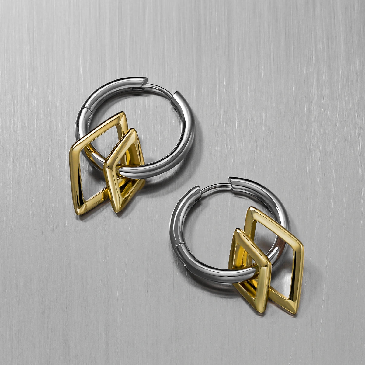 Two silver-tone hoop earrings with gold-tone geometric charms, angled view on a brushed metal surface