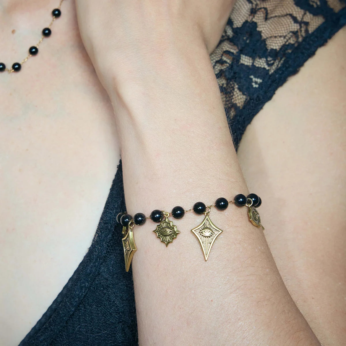A close-up of a person's wrist wearing a black beaded bracelet with antique bronze charms, including a star, an eye motif, and a crescent moon, styled with a dark lace top.
