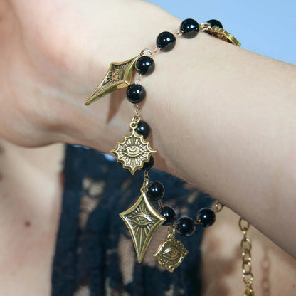 A close-up of a person's wrist wearing a beaded bracelet with black round beads and antique gold charms, including a diamond-shaped pendant with an eye motif and a small hanging eye charm.