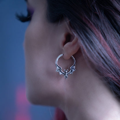Silver-tone bat-shaped hoop earring worn on ear, profile view against blurred background