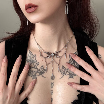 Model wearing a layered silver-tone necklace with spike charms and matching drop earrings over a tattooed chest