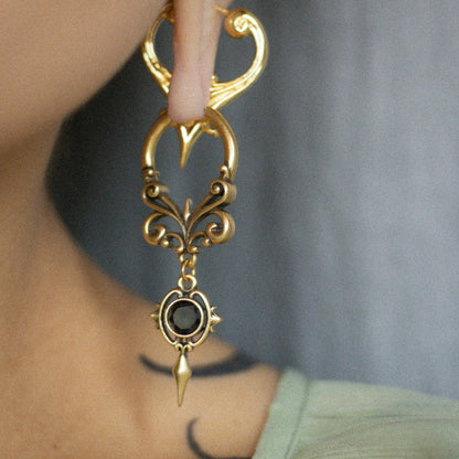 Close-up of a gold-tone Gothic Crest hoop earring with curled filigree and a black oval center stone