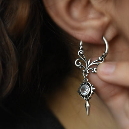 Close-up of silver-tone gothic crest earring with filigree design and clear crystal, held by fingers near ear