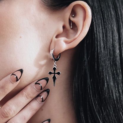 Close-up of a silver-tone hoop earring with a black cross charm worn on a model's ear