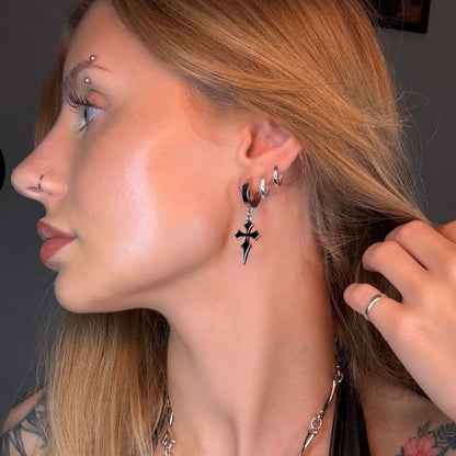 Model wears a silver-tone hoop earring with a black cross charm alongside a stacked ear piercing look