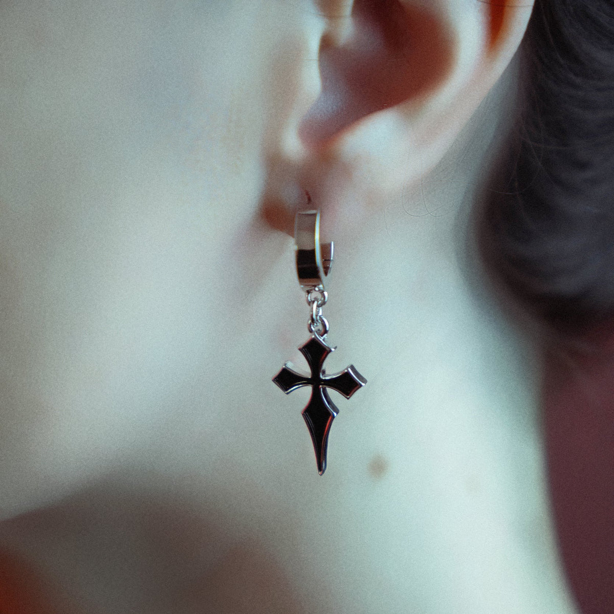 Side view of a black cross charm hanging from a silver-tone huggie hoop earring on a model