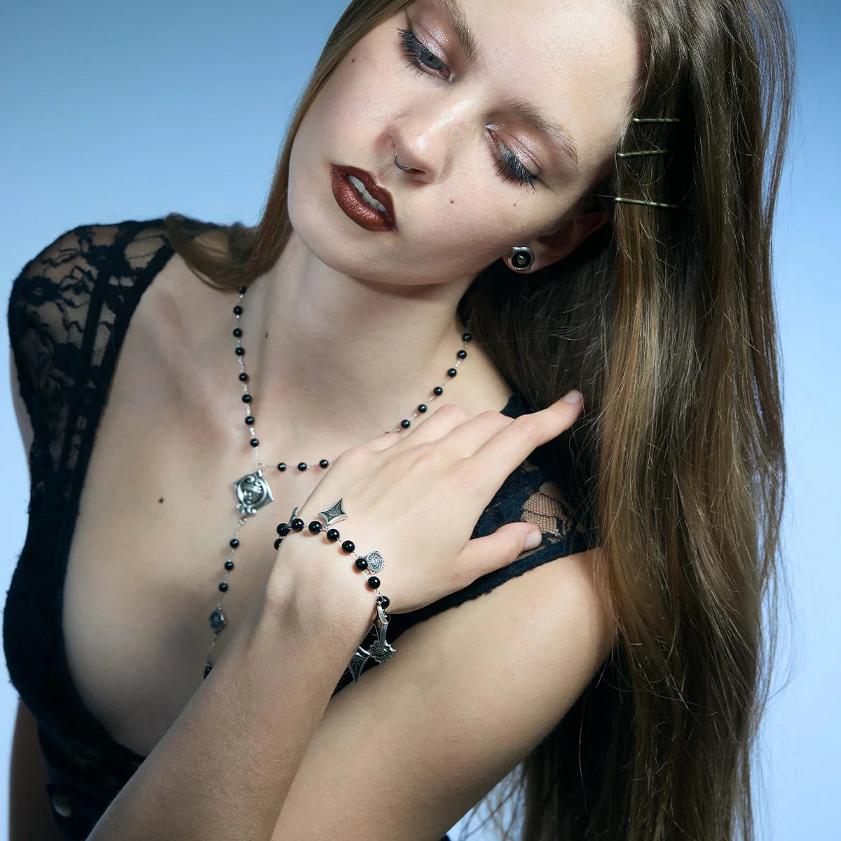 A woman with long brown hair models a gothic-style jewelry set featuring a silver and black beaded necklace, matching bracelet, and ring with dark red lipstick, set against a solid blue background.