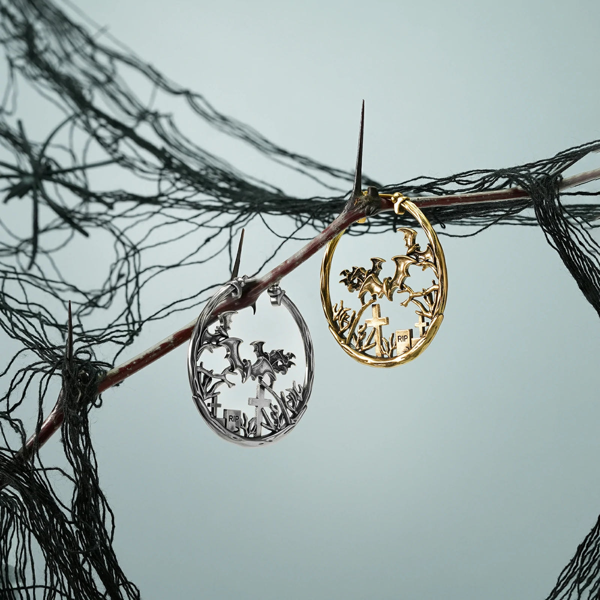 Silver and gold Graveyard Bat hoop earrings hanging from a dark branch