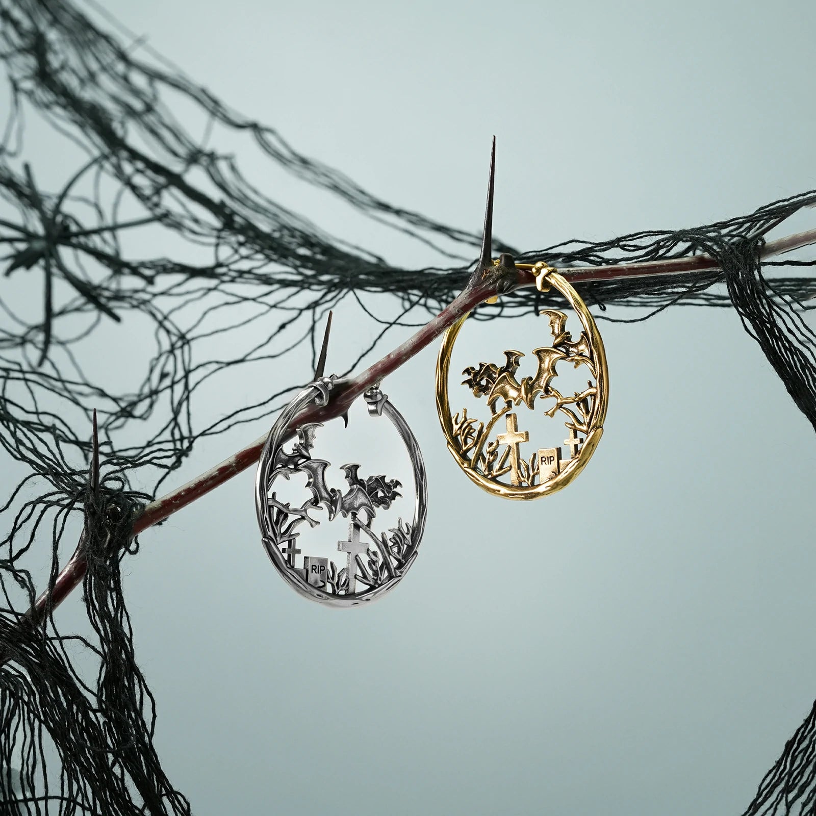 Silver and gold Graveyard Bat hoop earrings hanging from a dark branch