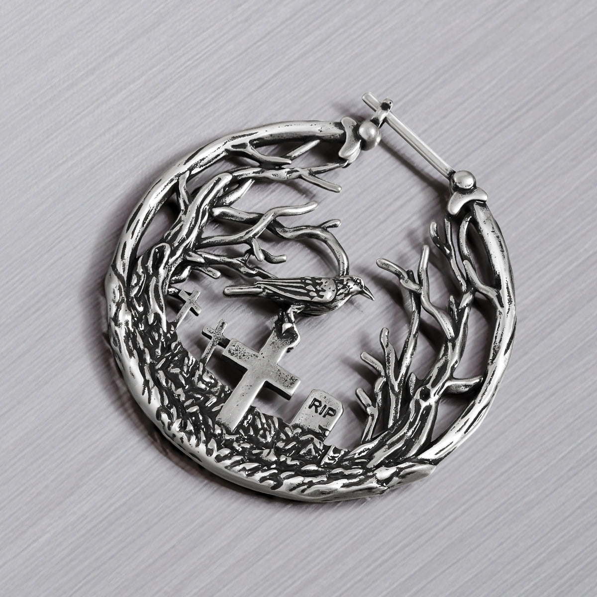 Close-up of a silver-tone hoop earring showing a raven perched above cross gravestones within a branch design