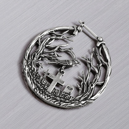 Close-up of a silver-tone hoop earring showing a raven perched above cross gravestones within a branch design