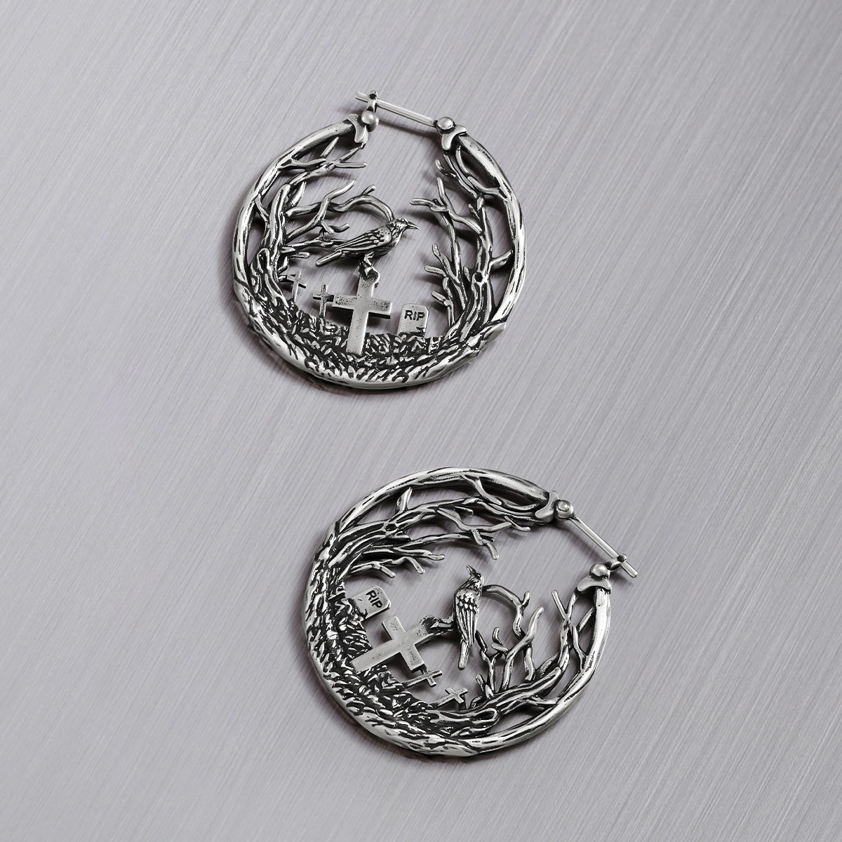 Pair of silver-tone hoop earrings with raven and cross gravestone details set within a branch motif, top view