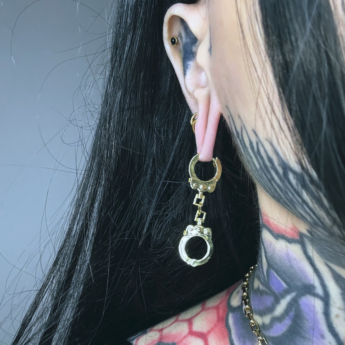 Close-up of a gold-tone handcuff chain earring worn on a model with dark hair and tattoos