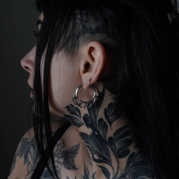 Model with dark hair and neck tattoos wearing a silver-tone hoop earring with wing-shaped detail, side profile