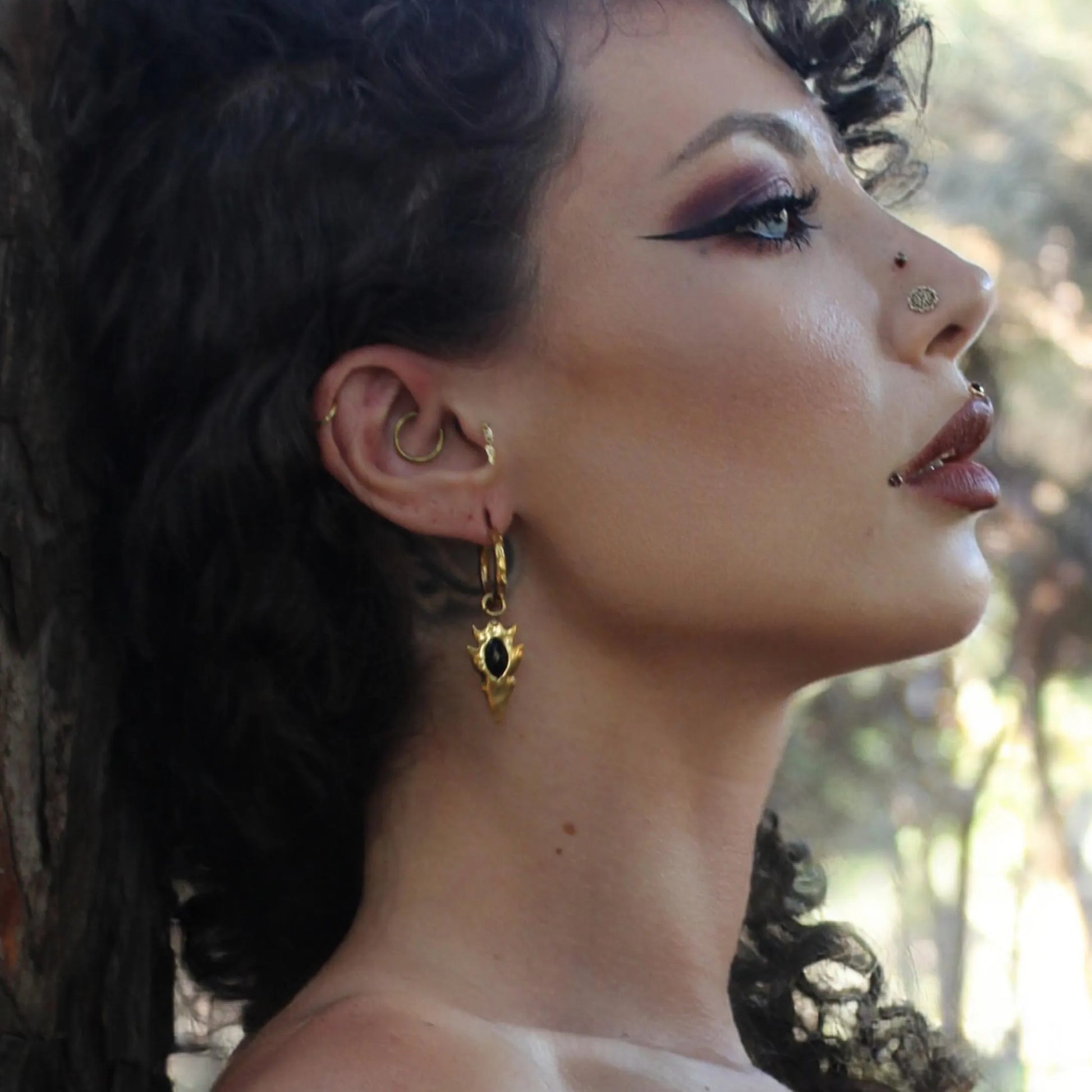 Model wearing the gold Knapped Stone Earrings with black obsidian arrowhead drops, hammered hoops, and dark forest-inspired styling.