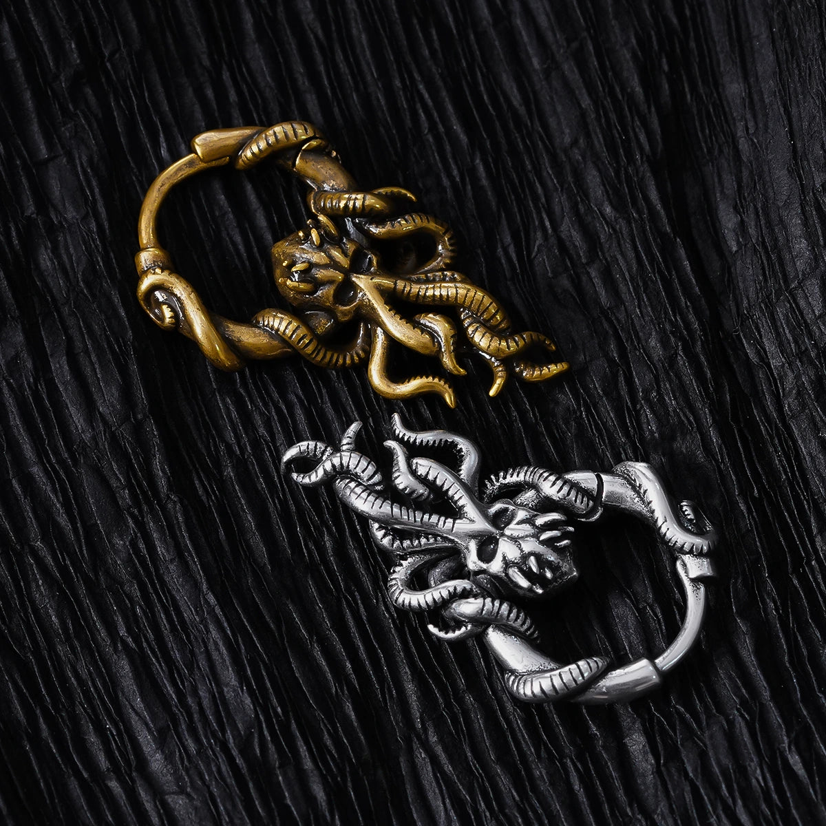 Gold-tone and silver-tone kraken hoop earrings laid on dark textured fabric