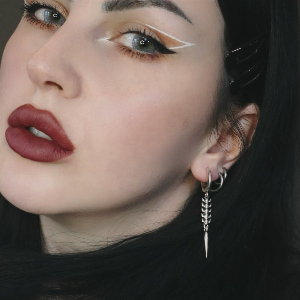 Longinus Abyss Stinger Earrings in 925 sterling silver, worn in a close brunette portrait with white graphic eyeliner and a b