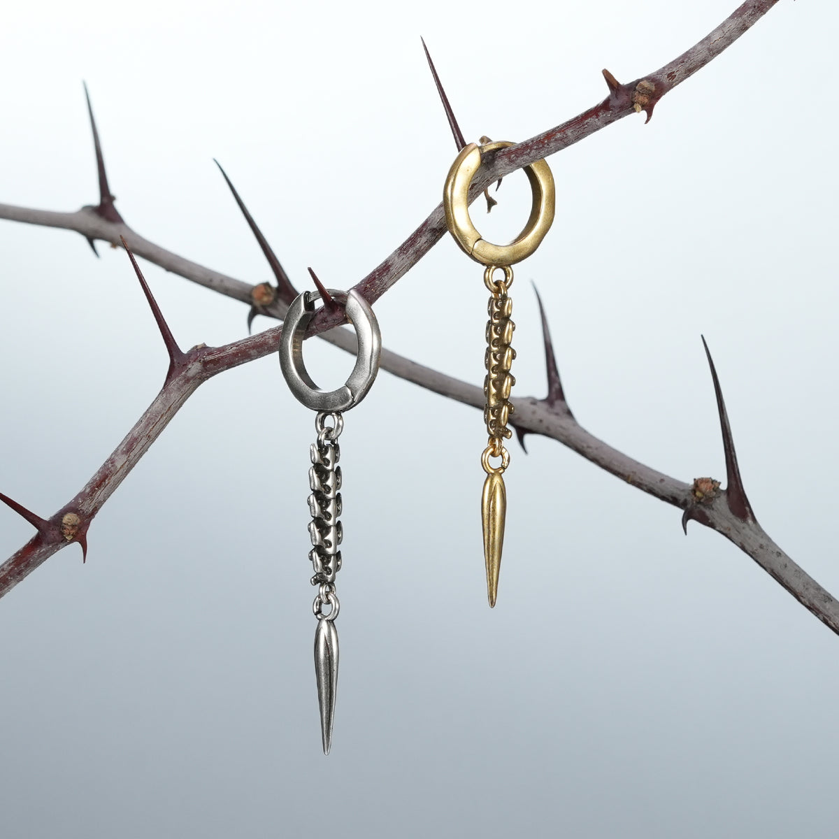 Silver and gold Longinus Abyss Stinger earrings hanging from a thorn branch