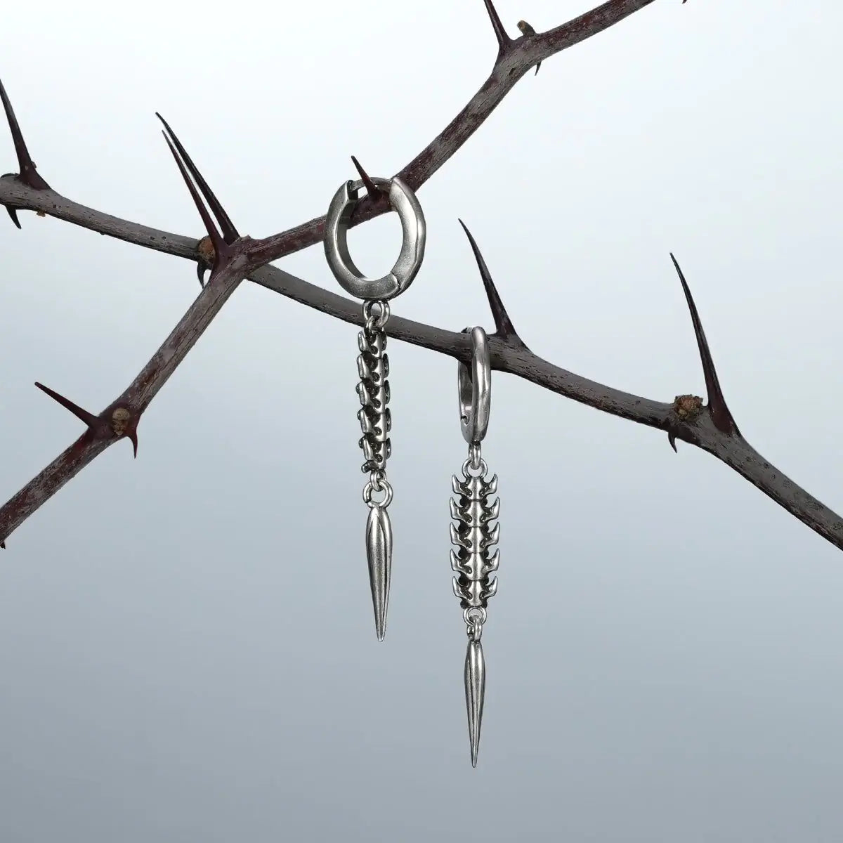 Silver spine and spike drop earrings hanging from a thorned branch against a light background