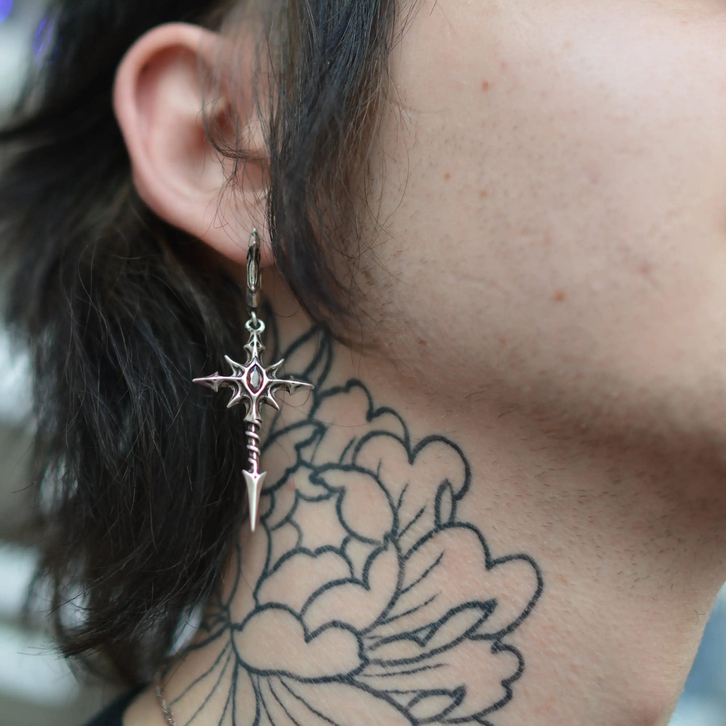 longinus blood relic earrings shown in a model close-up view, featuring a silver cross-shaped drop earring with a red gemstone detail worn on the ear