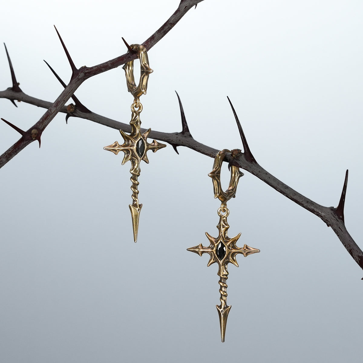 Silver and gold Longinus Blood Relic earrings displayed on a dark thorn branch