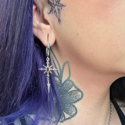 Silver-tone dagger cross earring with red gemstone worn by a model with purple hair and neck tattoo