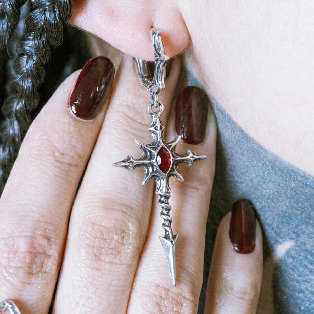 Close-up of a silver-tone dagger cross earring with red gemstone worn on an ear, held between fingers