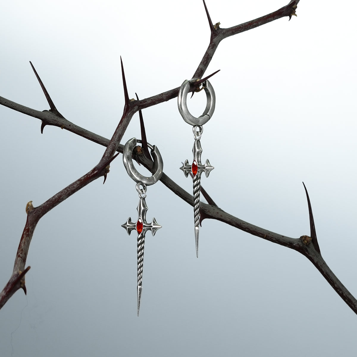 Two silver-tone Longinus Godpiercer sword hoop earrings hanging from dark thorn branches