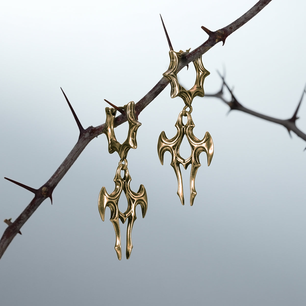 Pair of gold Longinus Judgment Talon earrings hanging from a thorn branch