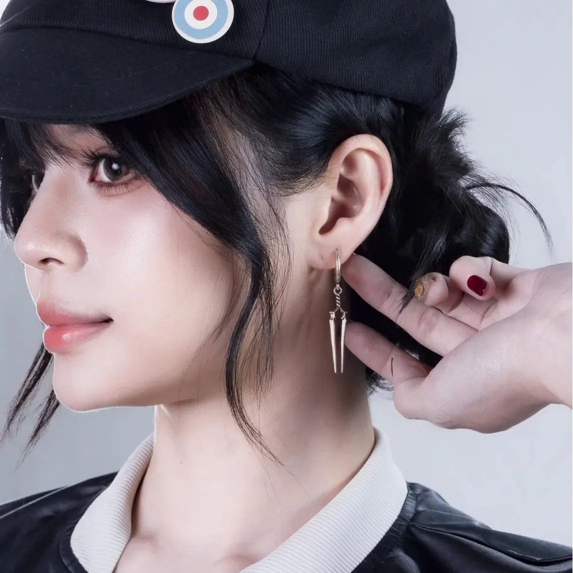 Longinus Twin Spear Earrings in 925 sterling silver, worn in side profile with black hair, a black cap, and a hand posed behi