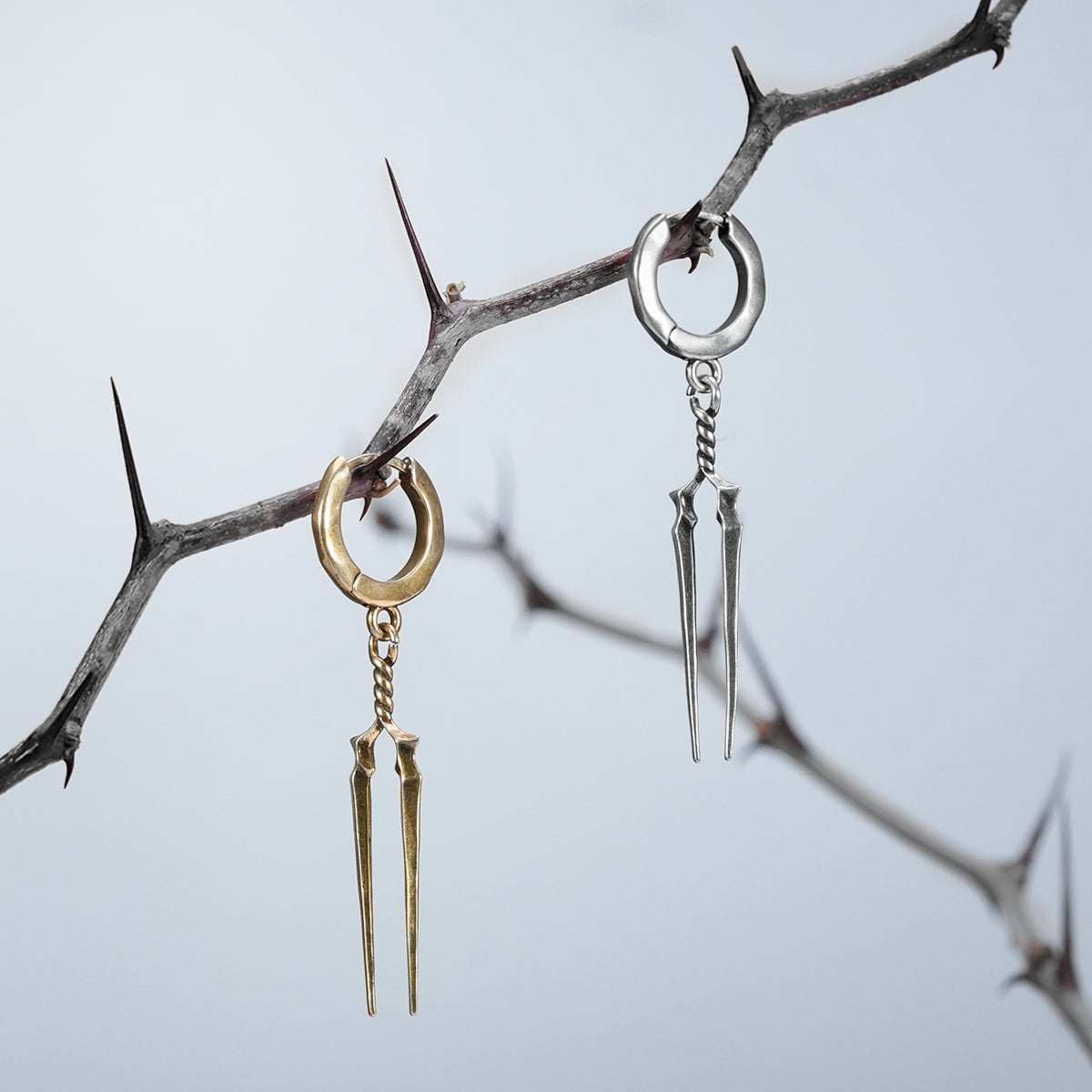 Gold and silver-tone Longinus Twin Spear hoop earrings hanging from a thorny branch outdoors