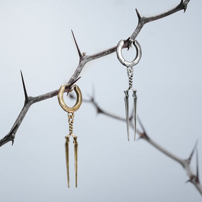 Gold and silver-tone Longinus Twin Spear hoop earrings hanging from a thorny branch outdoors