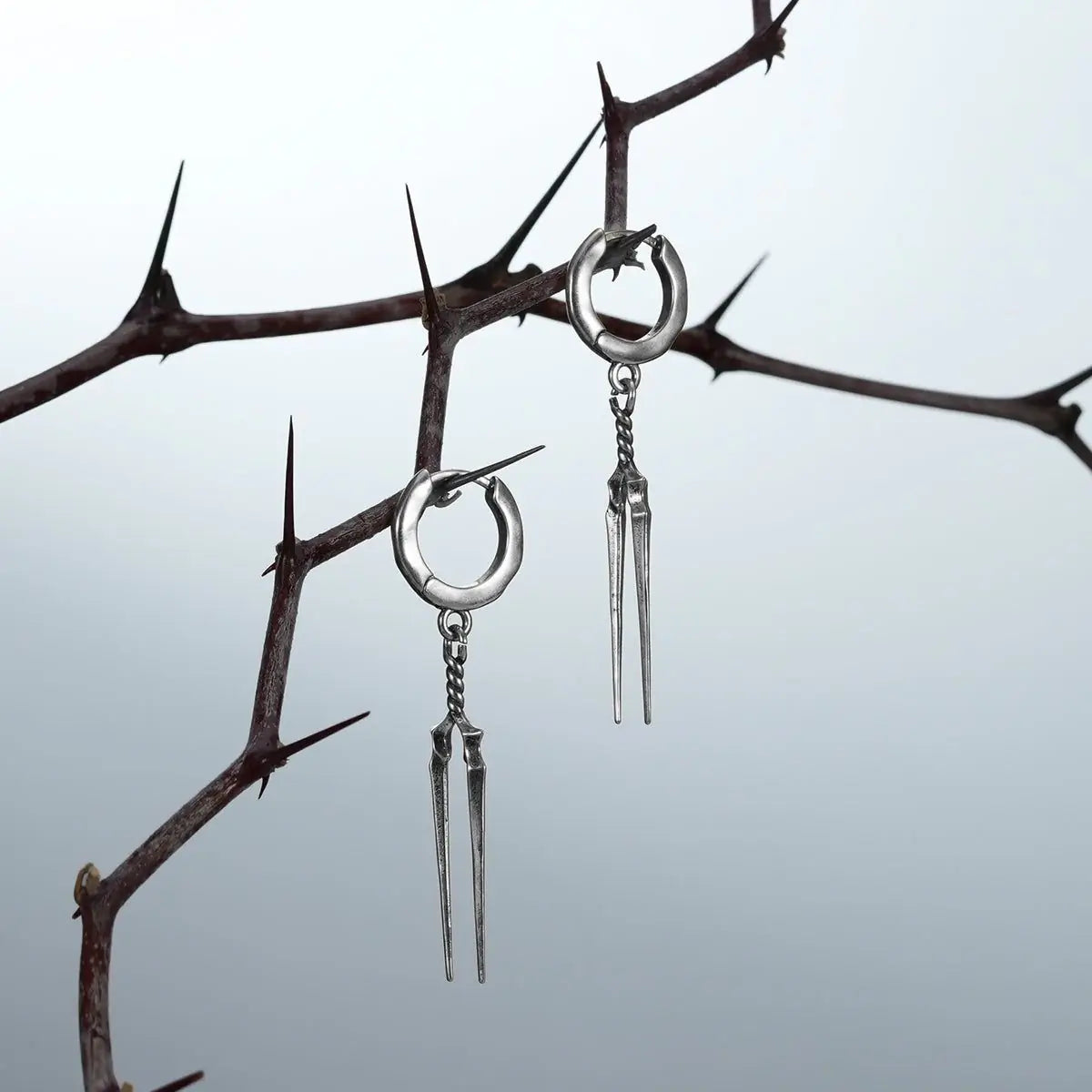 Two silver-tone Longinus Twin Spear hoop earrings hanging from dark thorn branches
