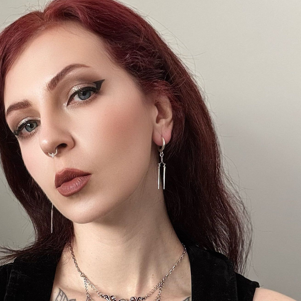 Model wearing a small silver-tone hoop earring with slender twin spear drops alongside a nose ring and necklace