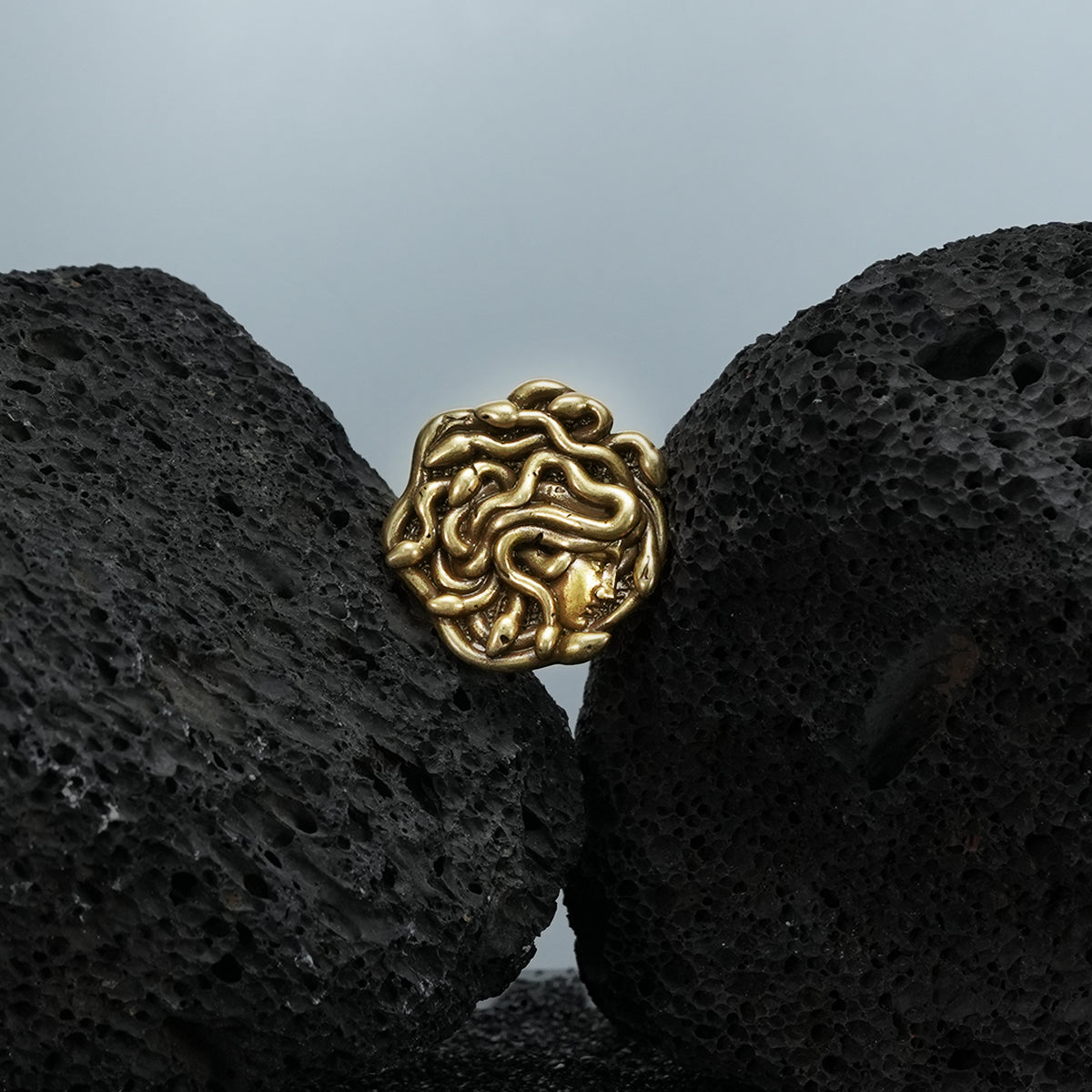 medusa unbound brooch in gold shown in a product view on black volcanic rock, featuring a sculpted medusa face formed by flowing snake details
