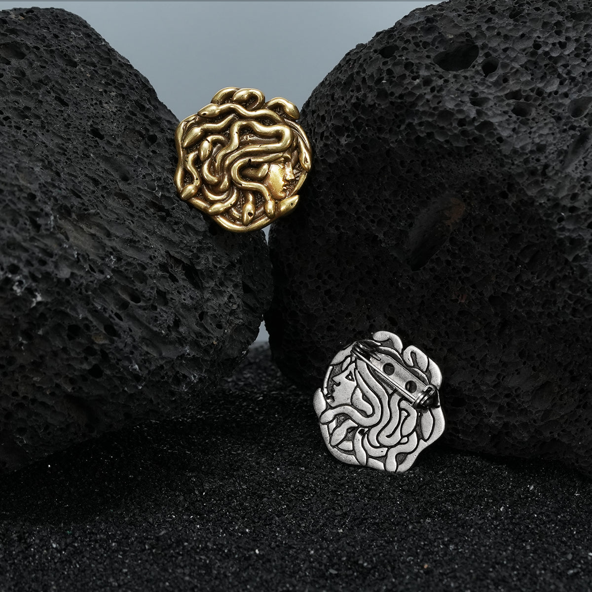 medusa unbound brooch shown in gold and silver in a product view on black volcanic rock, featuring a sculpted medusa face design