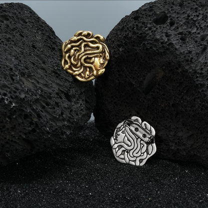 medusa unbound brooch shown in gold and silver in a product view on black volcanic rock, featuring a sculpted medusa face design