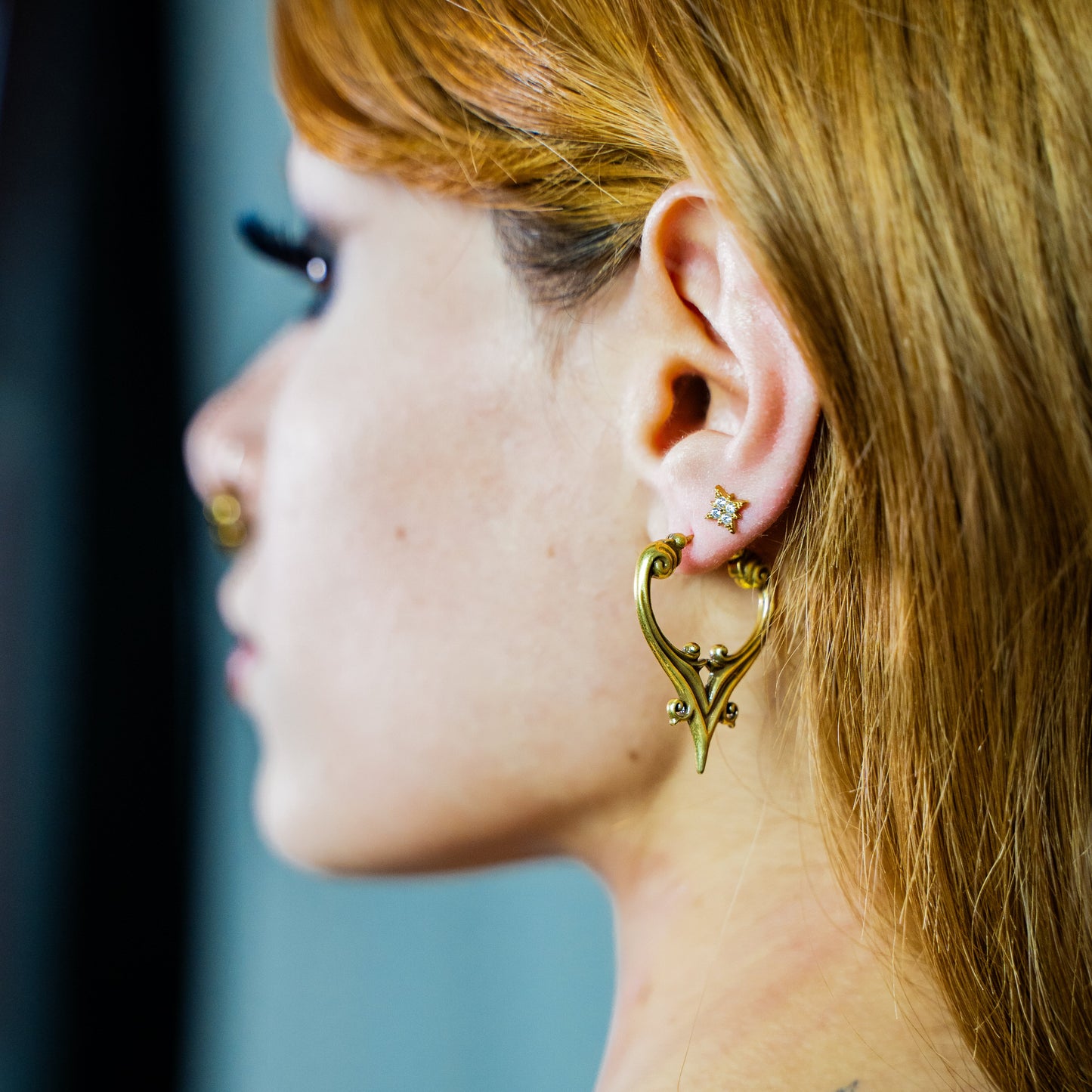 Side view of a gold-tone crest hoop earring worn alongside a stud, with a matching lobe earring visible in the background